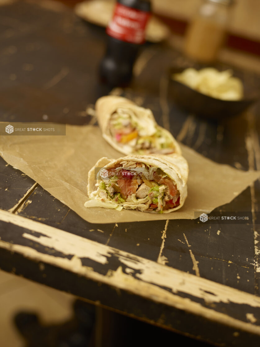 Chicken shawarma sandwich cut in half on brown parchment paper on a weathered table with a coke bottle and bowl of  chips in the background