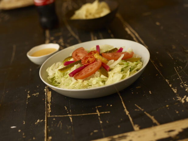 Salad in a round white bowl in foreground with dressing, soda in the background on a weathered table