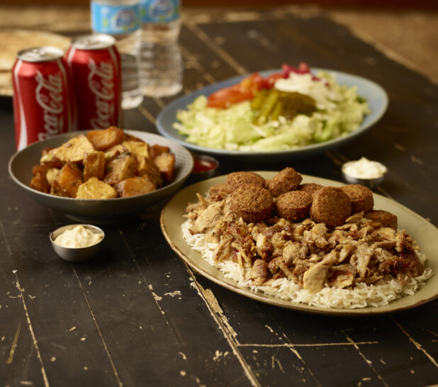 Shawarma and falafel on rice on a oval plate with a bowl of roasted potatoes, side salad, 2 cans of coke, 2 bottles of water and 2 dips in bowls in the background on a weathered table top
