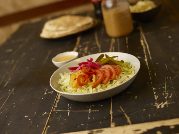 Middle eastern salad on a 45 degree angle with dressing, pitas, bowl of chips in the background on a weathered table