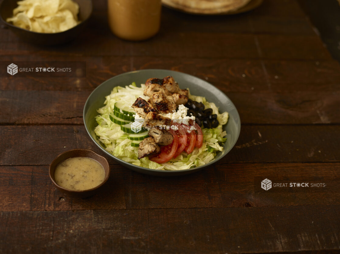 Greek salad with chicken in a bowl with a side bowl of vinaigrette dressing on a rustic table with potato chips in a side bowl in the background