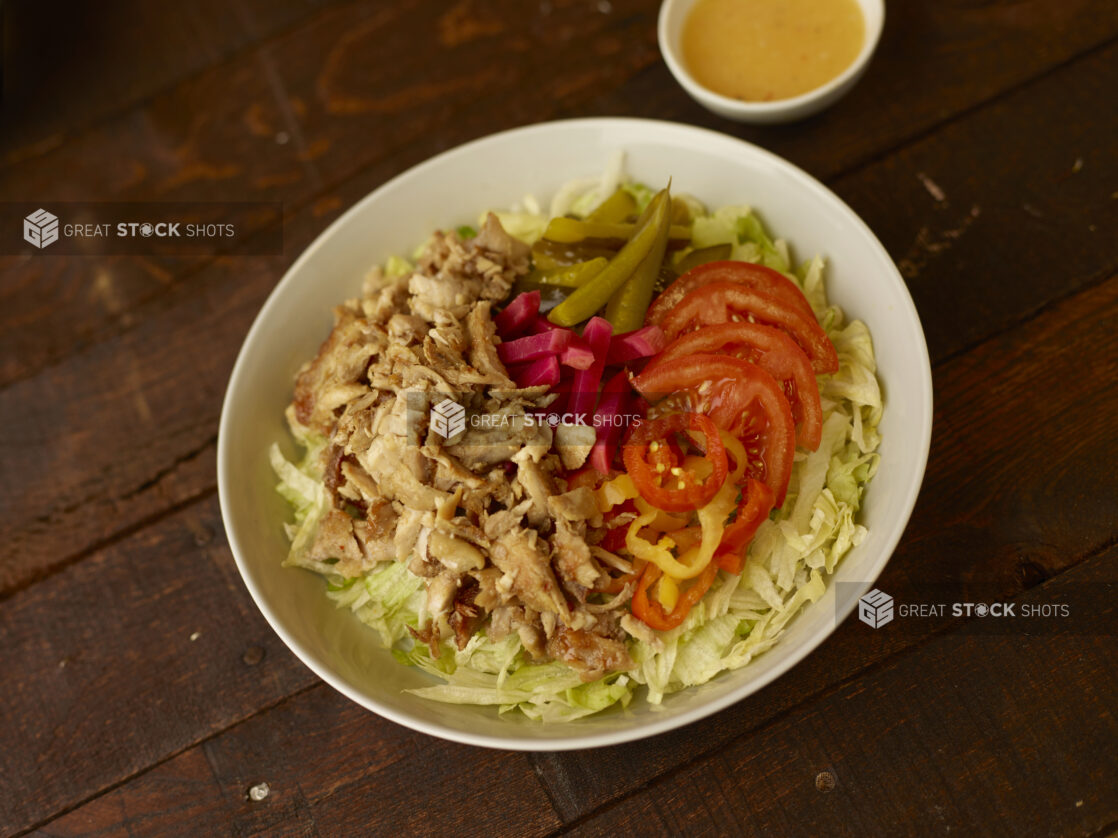 Overhead view of a chicken shawarma salad in a round white bowl with dressing on the side on a rustic wooden table