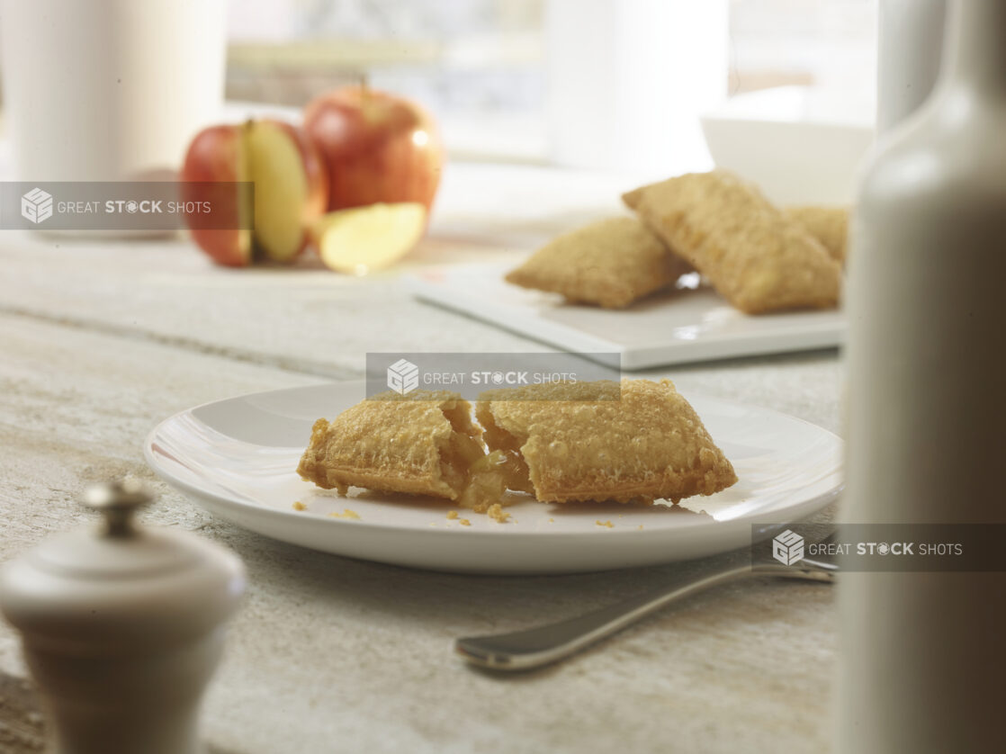 Hot apple pie on a round white dessert plate with more pies and apples in the background on a white wooden table