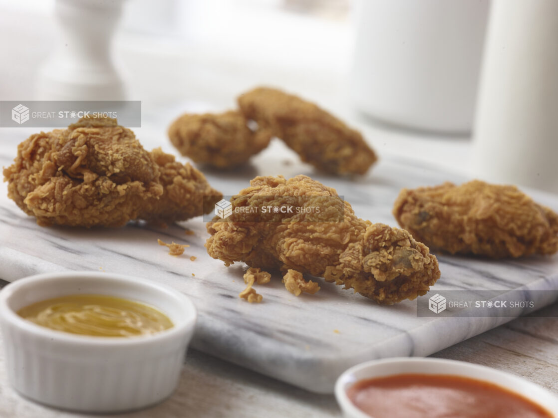 4 breaded  chicken wings displayed on a marble board with 2 dipping sauces in white ramekins, close up view