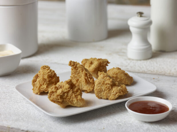 Fried chicken on a rectangular white platter with a ramekin of ketchup in the forefront with white accessories in the background