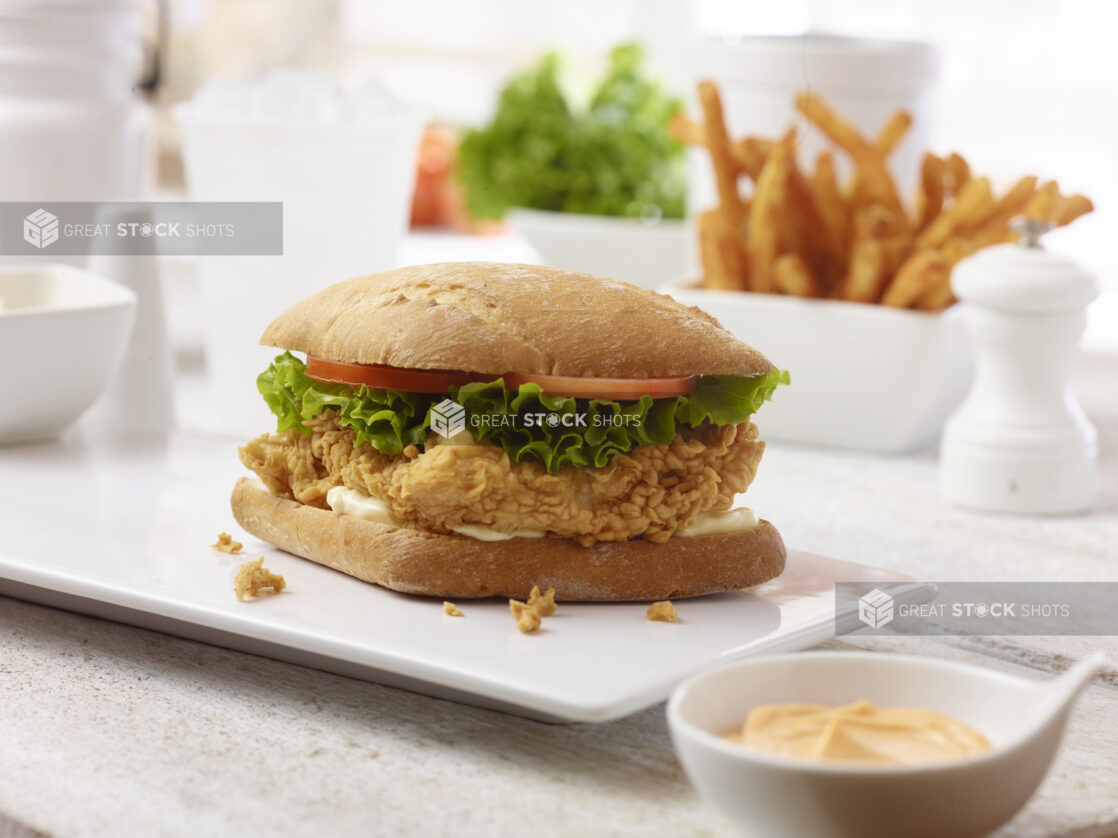 Crispy fried chicken sandwich with lettuce and tomato on a panini bun on a white platter with french fries in the background and a small bowl of chipotle mayo in the foreground