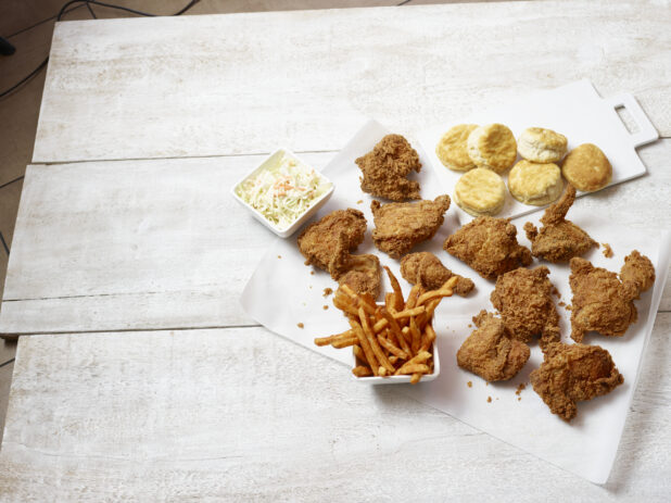 Overhead view of fried chicken on a white parchment paper with biscuits, french fries and coleslaw  on a white wooden table