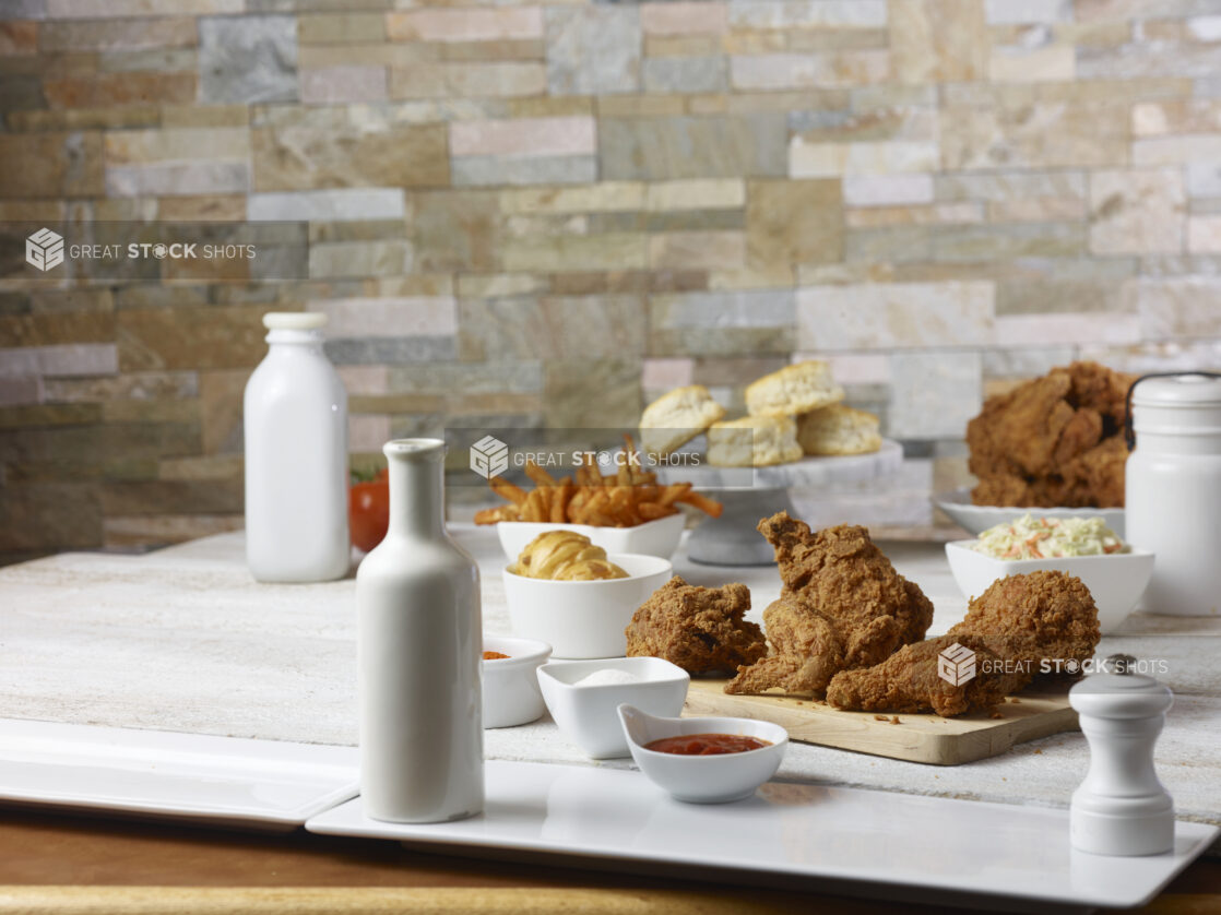 Fried chicken on a wooden cutting board with biscuits, french fries, whole tomatoes and coleslaw in the background with ramekins of ketchup, salt and seasoning in the foreground