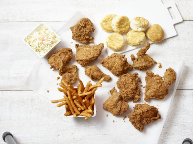 Fried chicken, seasoned french fries, biscuits and coleslaw on white parchment paper and white cutting board on a white wooden table