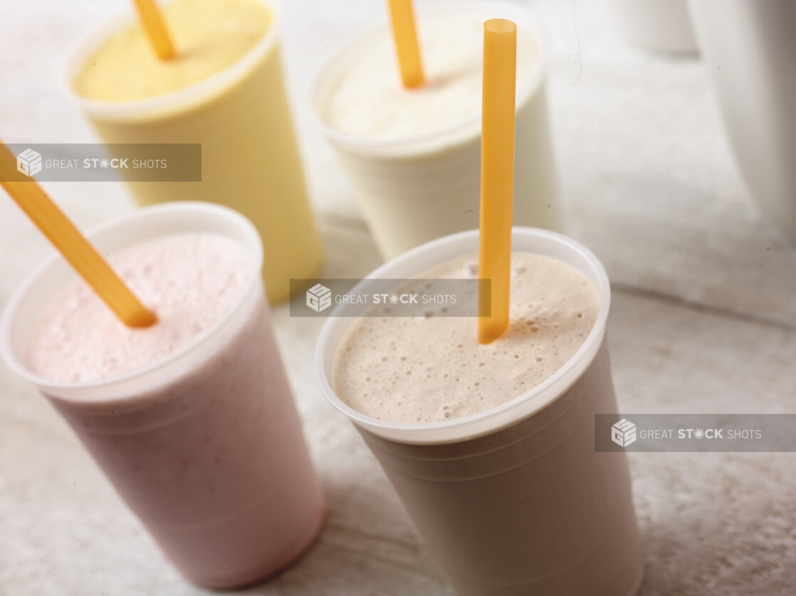 4 different flavours of smoothie/milkshake in plastic cups with orange straws, on an angle on a white wooden table