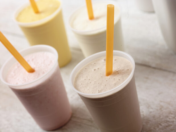 4 different flavours of smoothie/milkshake in plastic cups with orange straws, on an angle on a white wooden table
