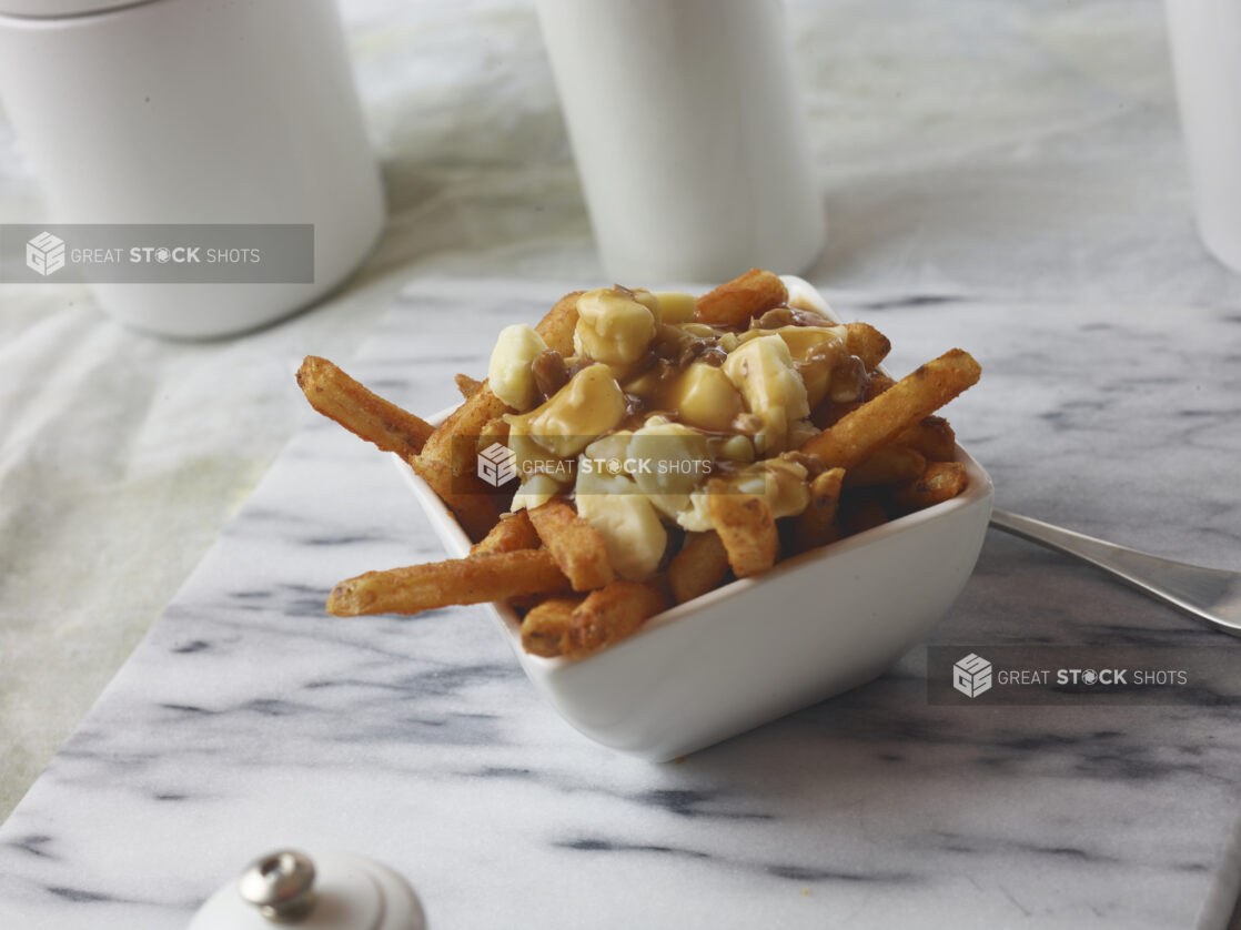 Poutine made with seasoned french fries with cheese curds and gravy on top in a small square white bowl on a marble cutting board