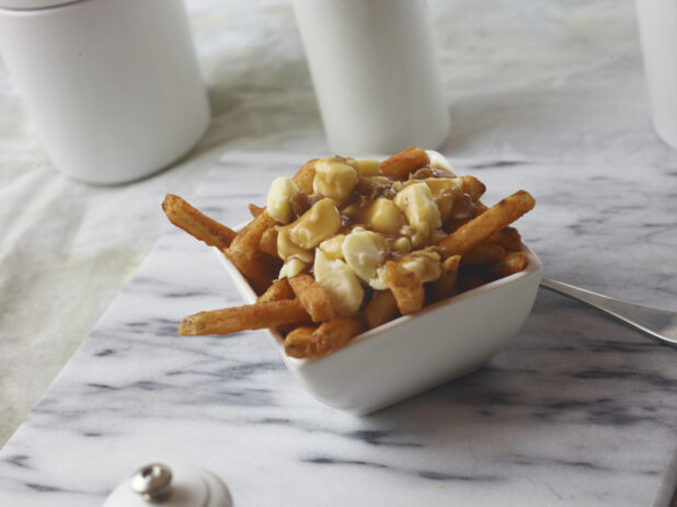 Poutine made with seasoned french fries with cheese curds and gravy on top in a small square white bowl on a marble cutting board