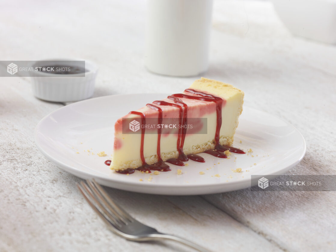 Strawberry cheesecake on a white dessert plate with a fork in the foreground on a white wooden table