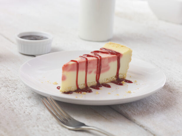 Strawberry cheesecake on a white dessert plate with a fork in the foreground on a white wooden table