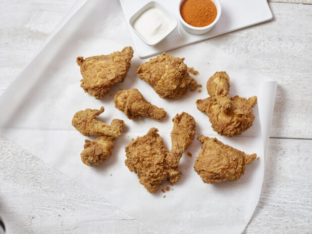 Overhead view of fried chicken on parchment paper on a white wooden table with a white ramekin of salt and a white ramekin of seasoning