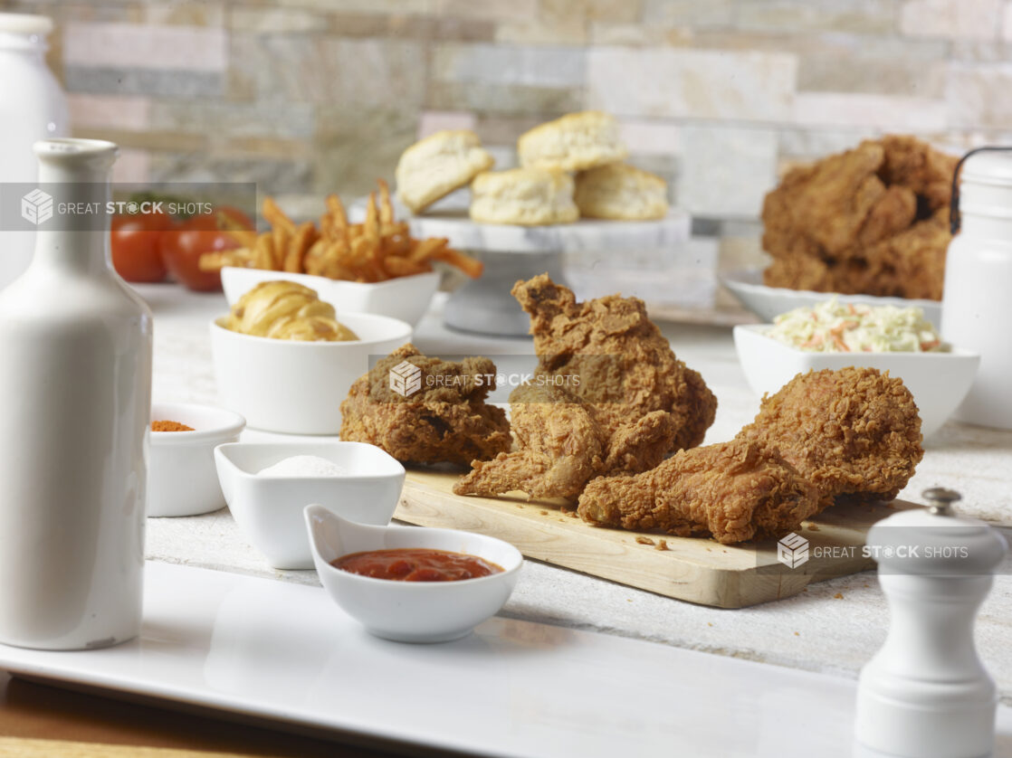 Fried chicken on a wooden cutting board, with fried chicken, biscuits, french fries, fresh tomatoes, coleslaw in the background with ketch, salt and seasoning in ramekins in the foreground