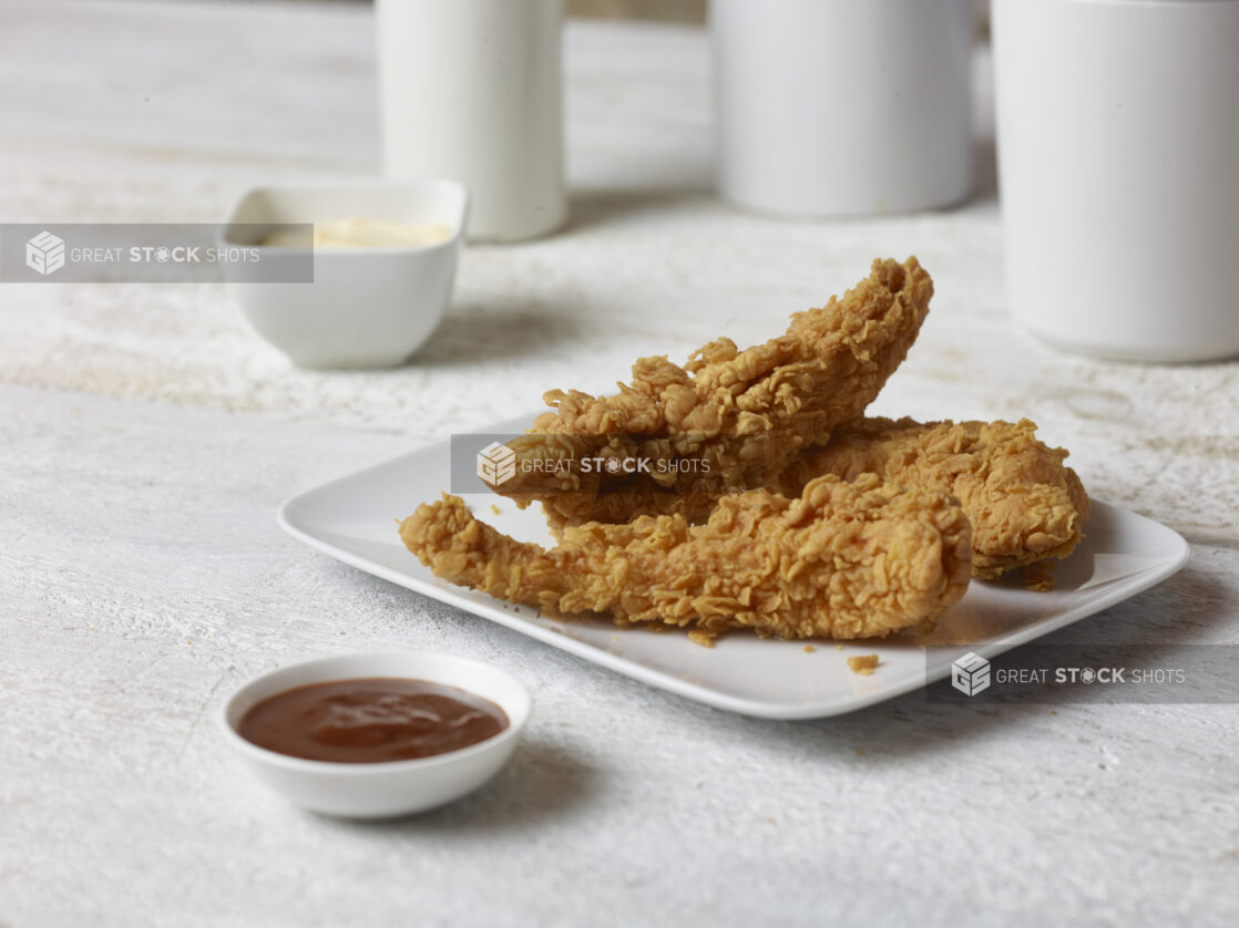 Crispy fried chicken strips on a square white plate with ketchup in a white ramekin in the foreground and chipotle mayo in a white ramekin in the background