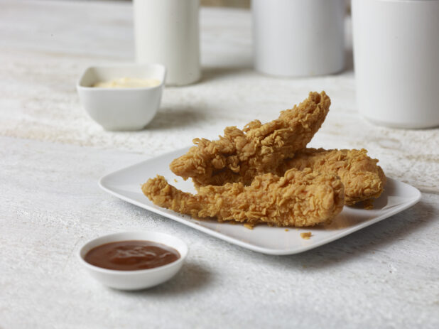 Crispy fried chicken strips on a square white plate with ketchup in a white ramekin in the foreground and chipotle mayo in a white ramekin in the background