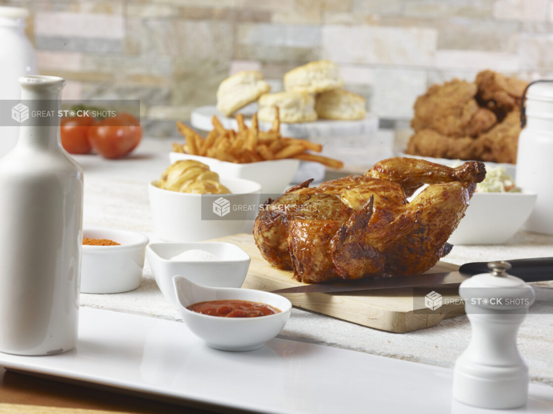 Roasted chicken on a wooden cutting board with biscuits, fried chicken, french fries, whole tomatoes and coleslaw in the background with ramekins of ketchup, salt and seasoning in the foreground