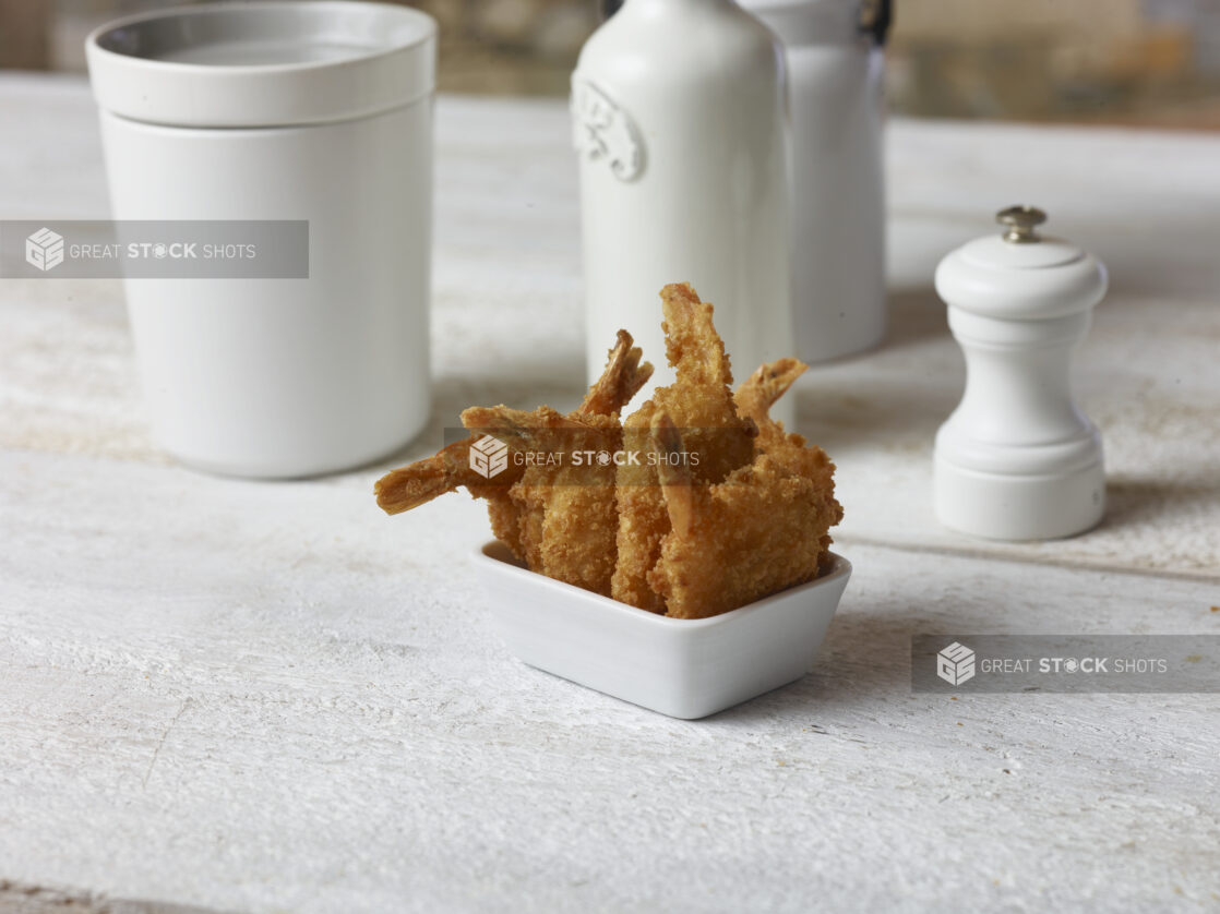 Breaded fried shrimp in a white square ramekin on a white wooden table surrounded by white accessories