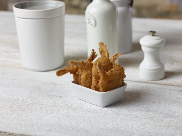 Breaded fried shrimp in a white square ramekin on a white wooden table surrounded by white accessories