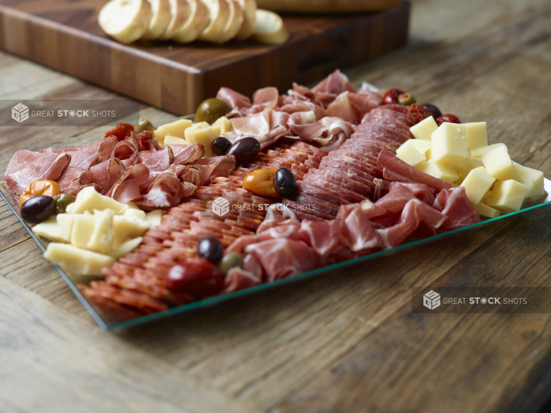 A charcuterie tray on a rectangular glass platter with sliced baguette on a wooden board in the background on a wooden table