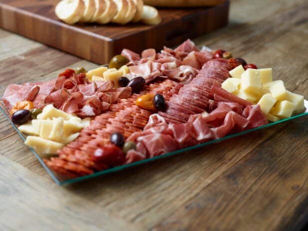 A charcuterie tray on a rectangular glass platter with sliced baguette on a wooden board in the background on a wooden table
