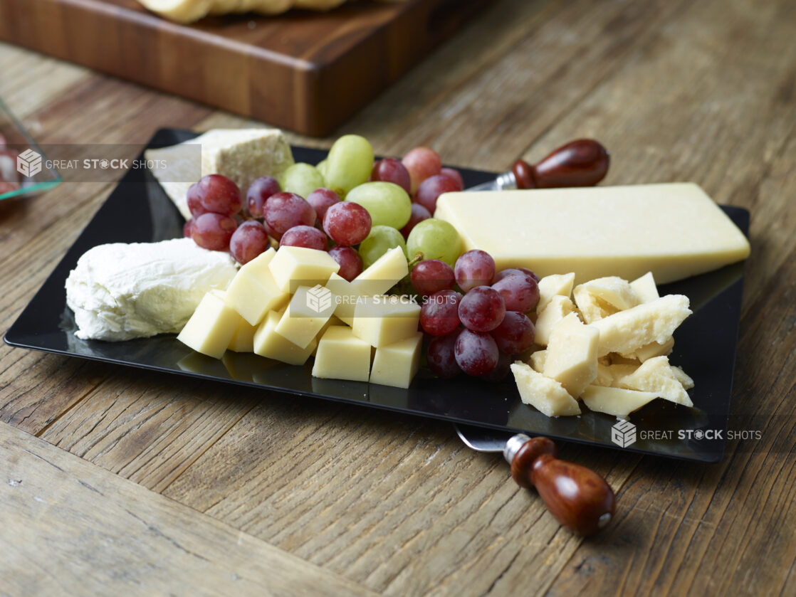 A cheese tray with various cheeses, red and green grapes on a black rectangular platter on a rustic wooden table