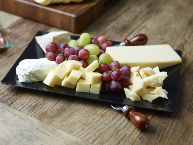 A cheese tray with various cheeses, red and green grapes on a black rectangular platter on a rustic wooden table