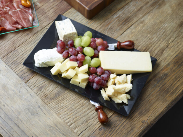 Overhead view of a cheese tray with various cheeses, red and green grapes on a black rectangular platter on a rustic wooden table