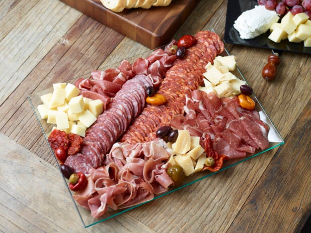 Overhead view of a charcuterie tray on a rectangular glass platter with a cheese tray in the background on a wooden table