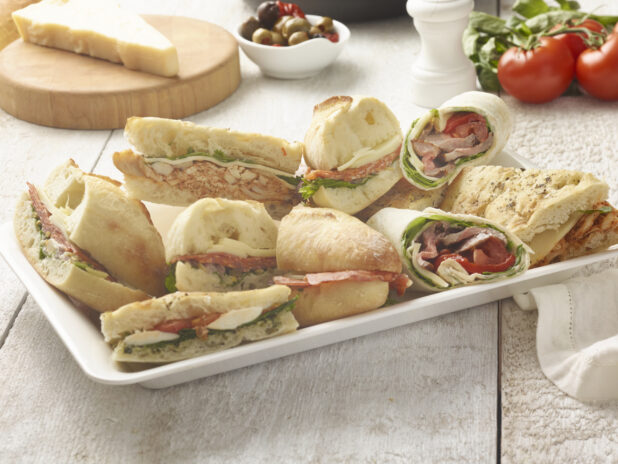 Hearty sandwiches on baguettes and focaccia on a rectangular white platter surrounded by fresh tomatoes, bowl of olives, parmesan cheese on a round wooden board