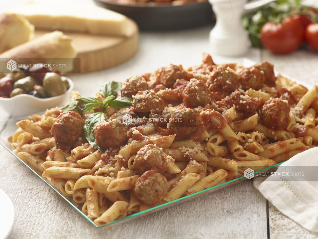 Large glass platter of penne pasta with meatballs and tomato sauce with basil garnish