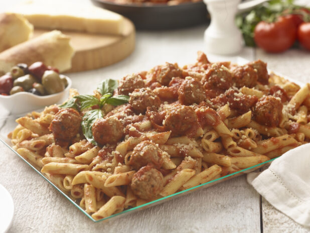 Large glass platter of penne pasta with meatballs and tomato sauce with basil garnish