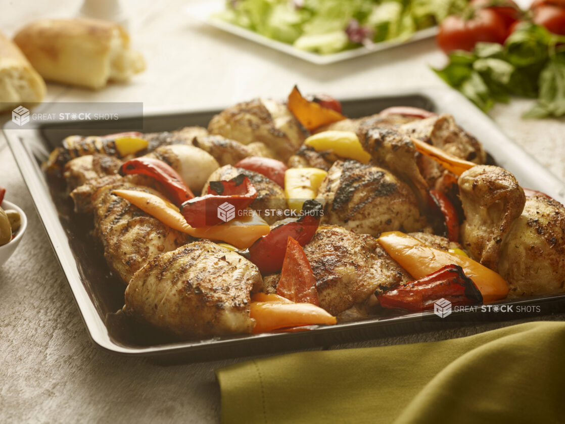 Roasted chicken and bell peppers on a square platter surrounded by fresh basil, salad and baguette