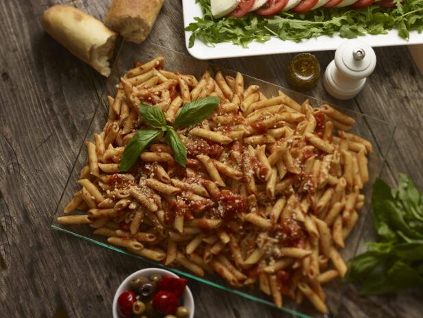 Overhead view of penne pasta in tomato sauce with a basil garnish surrounded by side dishes