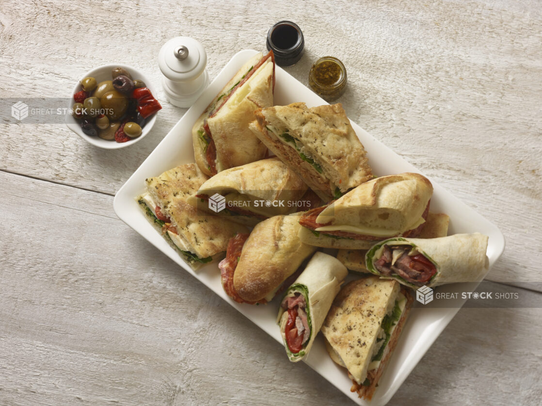 Hearty focaccia and baguette sandwiches on a white rectangular platter with a side bowl of olives