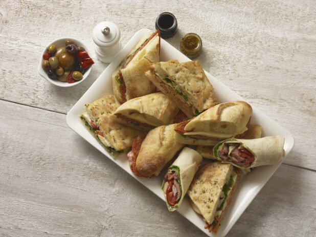 Hearty focaccia and baguette sandwiches on a white rectangular platter with a side bowl of olives