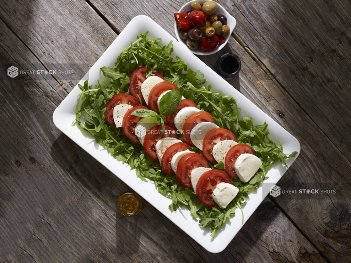 Caprese salad on a bed of arugula on a white platter with olives and dressing on the side on a rustic wooden table