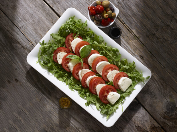 Caprese salad on a bed of arugula on a white platter with olives and dressing on the side on a rustic wooden table