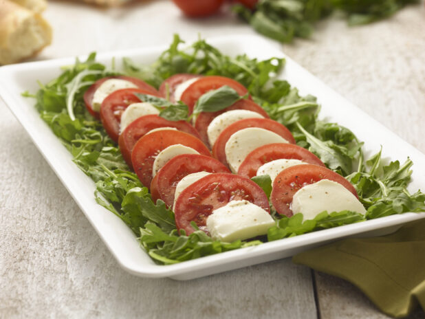 Family-style caprese salad on arugula, undressed, on a white platter on a whitewashed wood surface