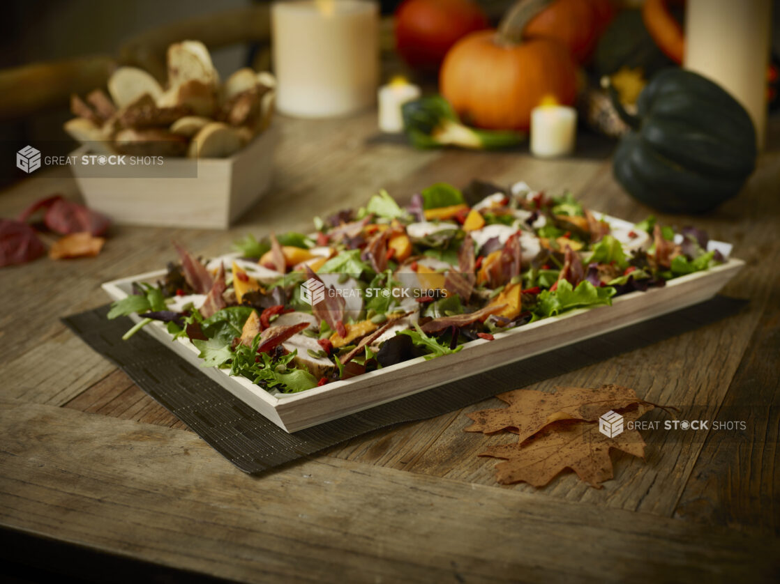 Fall salad on a wood tray with fall decorations/setting and a wood box with crusty sliced bread all on a rustic wooden table