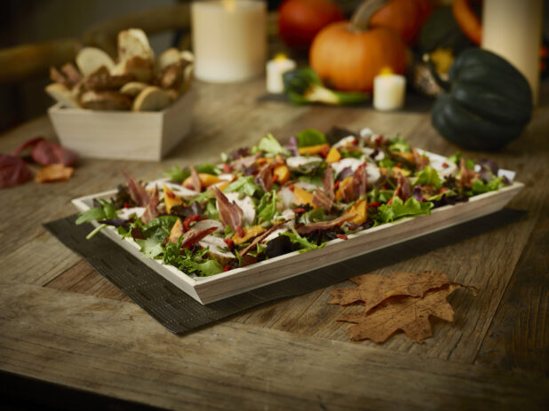 Fall salad on a wood tray with fall decorations/setting and a wood box with crusty sliced bread all on a rustic wooden table