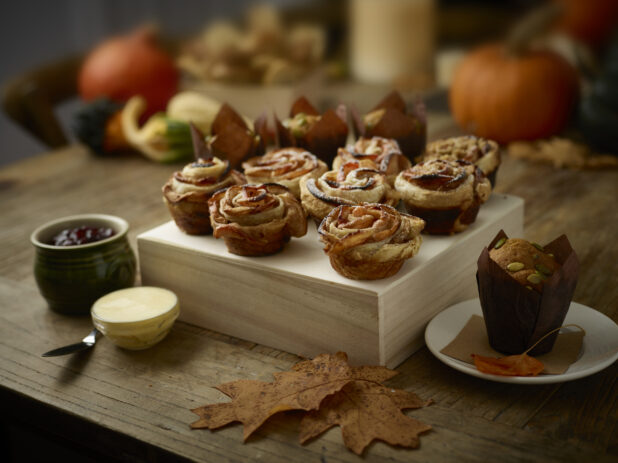 Apple rose tarts on a wood box with jam and butter in the foreground, pumpkin seed muffin on the side with a festive fall-themed background
