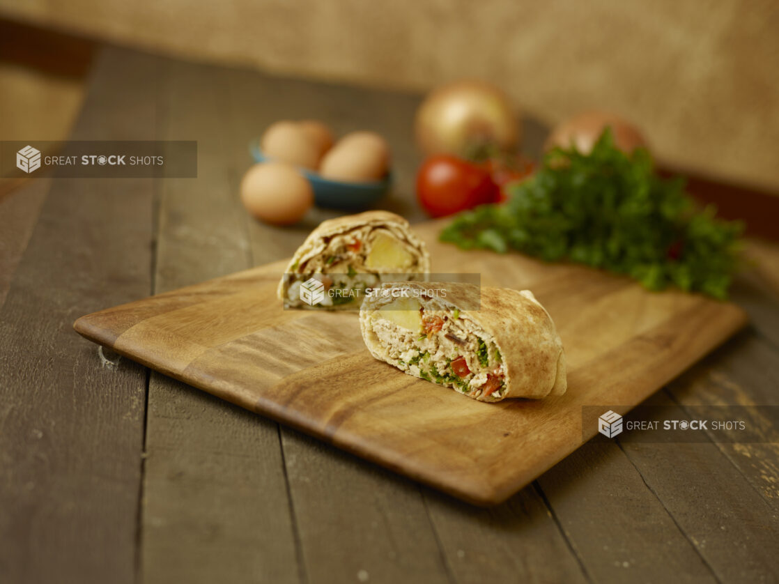 Breakfast egg wrap on a wooden cutting board, with wholes eggs, parsley, tomatoes and onions in the background all on a rustic wooden table