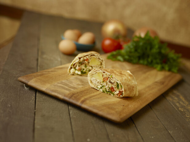 Breakfast egg wrap on a wooden cutting board, with wholes eggs, parsley, tomatoes and onions in the background all on a rustic wooden table