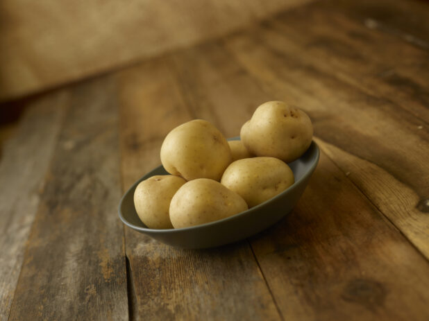 A bowl of whole/uncooked potatoes on a rustic wooden table on a 45 degree angle