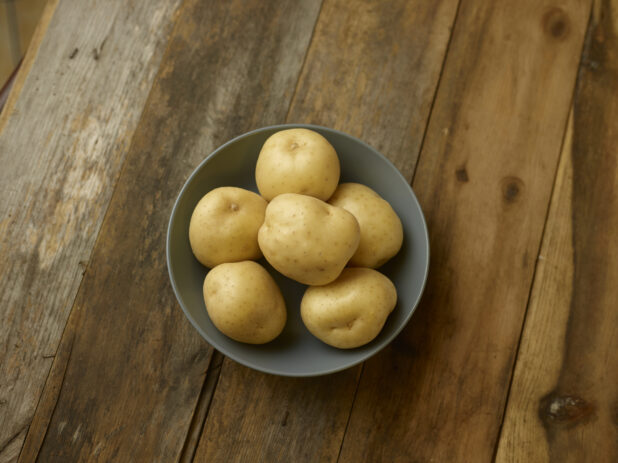 A bowl of whole/uncooked potatoes on a rustic wooden table