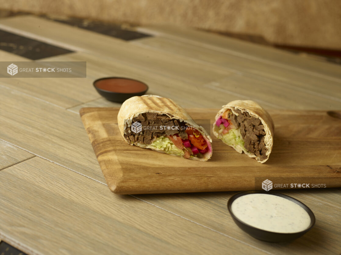 Beef shawarma on a wooden board cut in half with dips on the side on a 45 degree angle